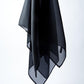 A sheer black silk scarf draped vertically on a white background. The photograph emphasizes the fabric's fine texture, transparency, and elegant drape, showcasing the garment as a minimalist fashion accessory.