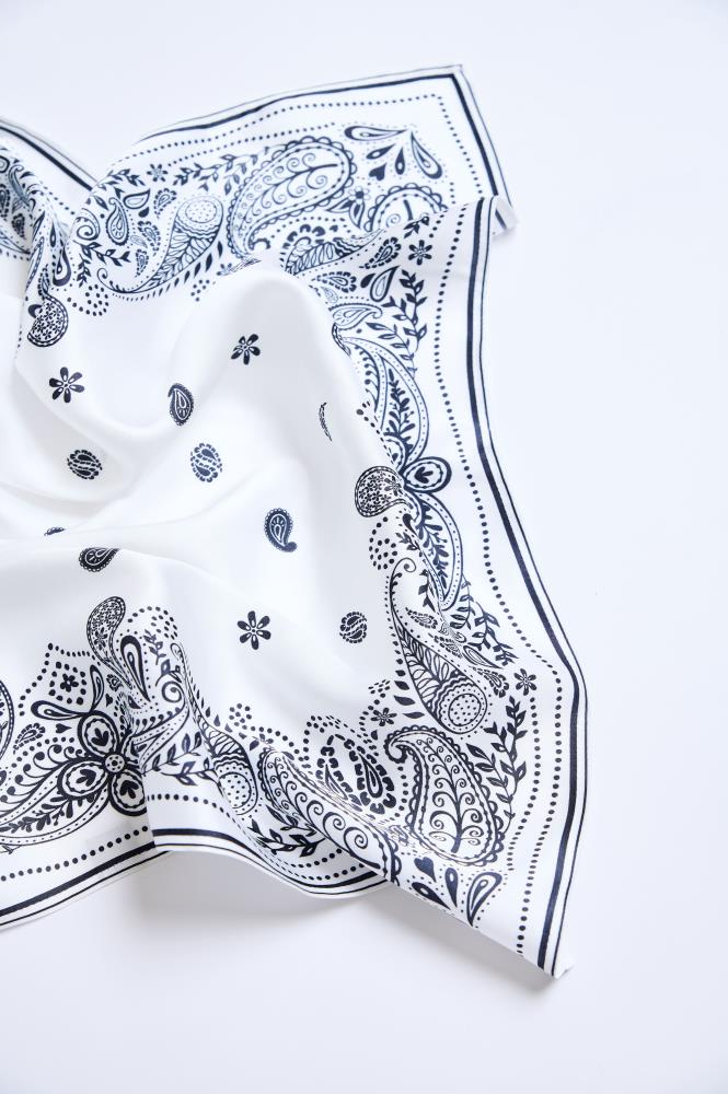 A crumpled white scarf with a black pattern, laid out on a light background. The close-up shot highlights the fabric's folds, adding depth to the image and emphasizing the material's softness. The accessory is decorated with a classic paisley motif and small floral elements