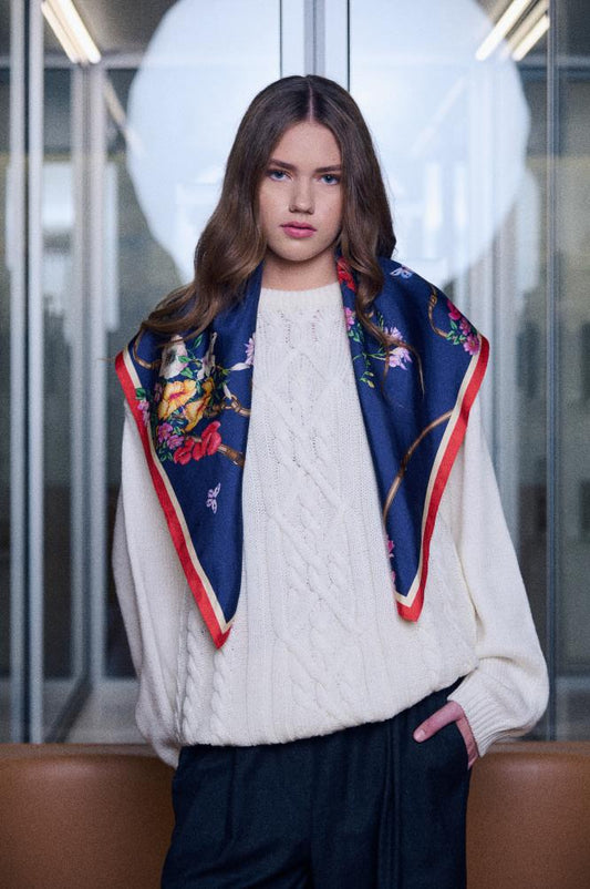 A woman with long wavy hair looks at the camera, wearing a light-colored cable-knit sweater and dark trousers. A luxurious navy blue silk scarf with a vibrant floral pattern and a red border is draped over her shoulders. The ends of the scarf hang symmetrically in front, forming a triangle and adding elegance to the look. The background is neutral and bright, keeping all the focus on the model and the accessory.
