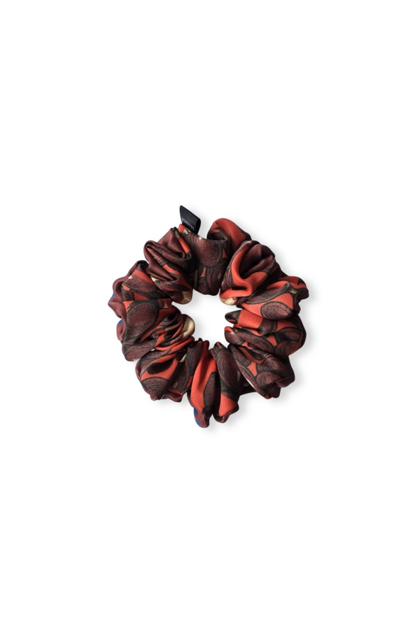A top-down view of a luxury patterned silk scrunchie, displayed flat on a white background. It features a deep reddish-orange and dark brown abstract pattern with a small brand tag.