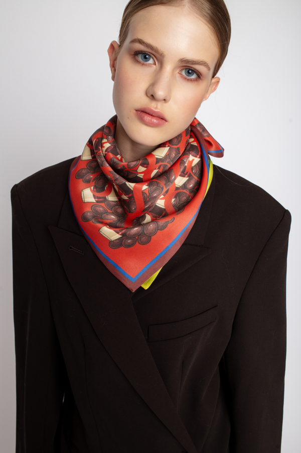A portrait focusing on a woman wearing a square silk scarf tied at her neck in triangle. The scarf has a dynamic pattern with prominent red and chocolate-brown circular motifs and a yellow corner, paired with a brown blazer.