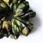 A detailed close-up of a luxury olive-patterned silk scrunchie, showcasing vibrant green and yellow whole olives on gathered fabric folds. 