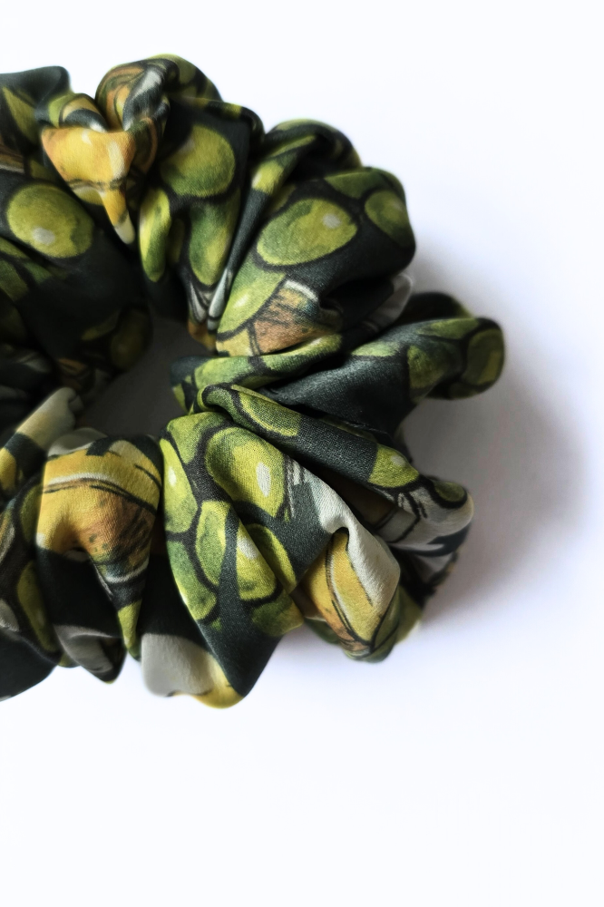 A detailed close-up of a luxury olive-patterned silk scrunchie, showcasing vibrant green and yellow whole olives on gathered fabric folds. 