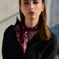 A model poses in a fashionable look with a  silk scarf stylishly tied around her neck. The scarf features a vibrant black and pink graphic pattern that stands out in contrast against a dark shirt. Natural lighting and a minimalist setting emphasize the modern aesthetic and elegant style of the accessory.