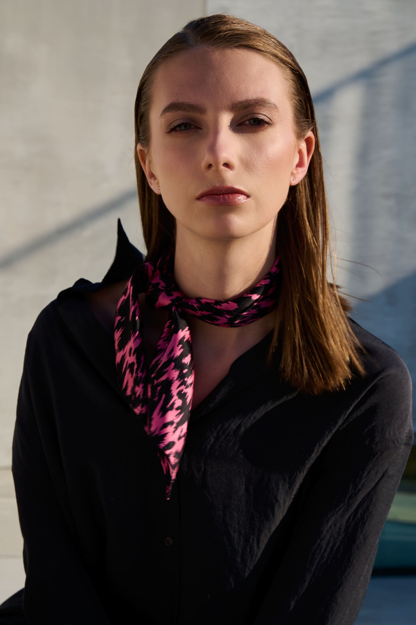 A model poses in a fashionable look with a  silk scarf stylishly tied around her neck. The scarf features a vibrant black and pink graphic pattern that stands out in contrast against a dark shirt. Natural lighting and a minimalist setting emphasize the modern aesthetic and elegant style of the accessory.