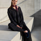 A model sits on concrete stairs in an industrial setting, wearing a loose black suit and a vibrant silk "Twilly" scarf. The scarf is black and pink with a graphic pattern, stylishly tied around her neck, adding a colorful accent to the minimalist look. Blue velvet shoes and sun shadows complement the modern and elegant composition of the photo.