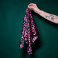 A hand with a tattoo holding a vibrant black and pink patterned silk scarf against a dark green background. The photo highlights the fabric's sheen, dynamic graphic pattern, and high-quality texture. A stylish and bold accessory that adds a modern accent to any look.