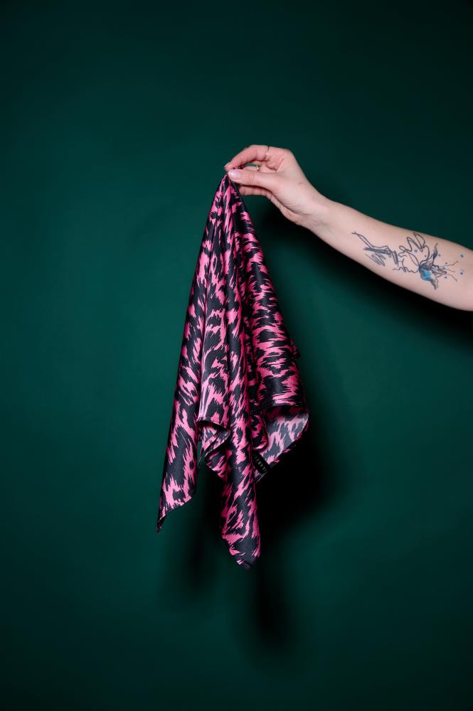 A hand with a tattoo holding a vibrant black and pink patterned silk scarf against a dark green background. The photo highlights the fabric's sheen, dynamic graphic pattern, and high-quality texture. A stylish and bold accessory that adds a modern accent to any look.