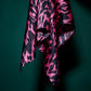 A detail of a vibrant pink and black patterned silk scarf against a dark green background. The photo shows the aesthetic drape of the fabric, a soft silk sheen, and a dynamic, contrasting graphic pattern. This composition emphasizes the accessory's luxury, modern design, and high quality.