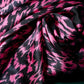 A detail of a luxurious silk scarf featuring a vibrant pink and black graphic pattern. The fabric is draped to highlight its soft sheen, supple texture, and dynamic design. A dark green background creates a stylish contrast, emphasizing the modern aesthetic and high quality of the accessory.