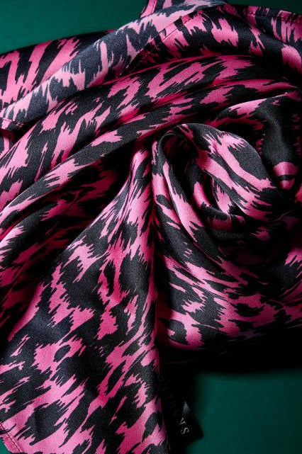 A detail of a luxurious silk scarf featuring a vibrant pink and black graphic pattern. The fabric is draped to highlight its soft sheen, supple texture, and dynamic design. A dark green background creates a stylish contrast, emphasizing the modern aesthetic and high quality of the accessory.