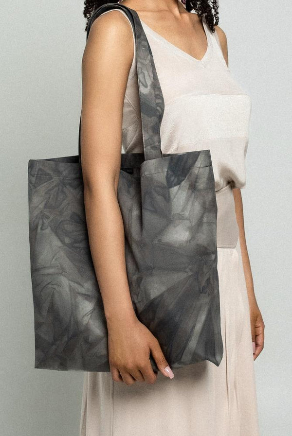 A close-up studio portrait of a woman model with dark, curly hair wearing a light beige, textured linen dress and holding a large grey and black tie-dye printed canvas tote bag over her shoulder.
