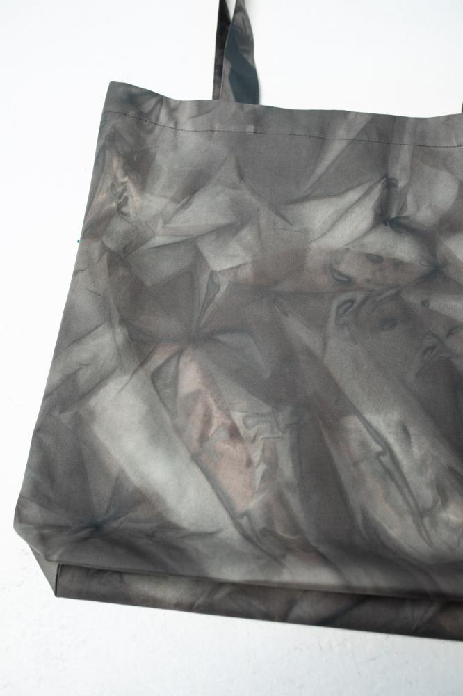 A close-up detail shot focusing on the abstract, smoky tie-dye pattern on a grey canvas tote bag, showing hints of brown. Displayed against a clean white background.