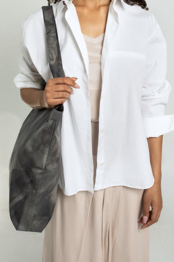 A close-up studio portrait of a woman wearing a white unbuttoned shirt over a beige dress, holding a dark grey and black tie-dye canvas tote bag by its long strap.