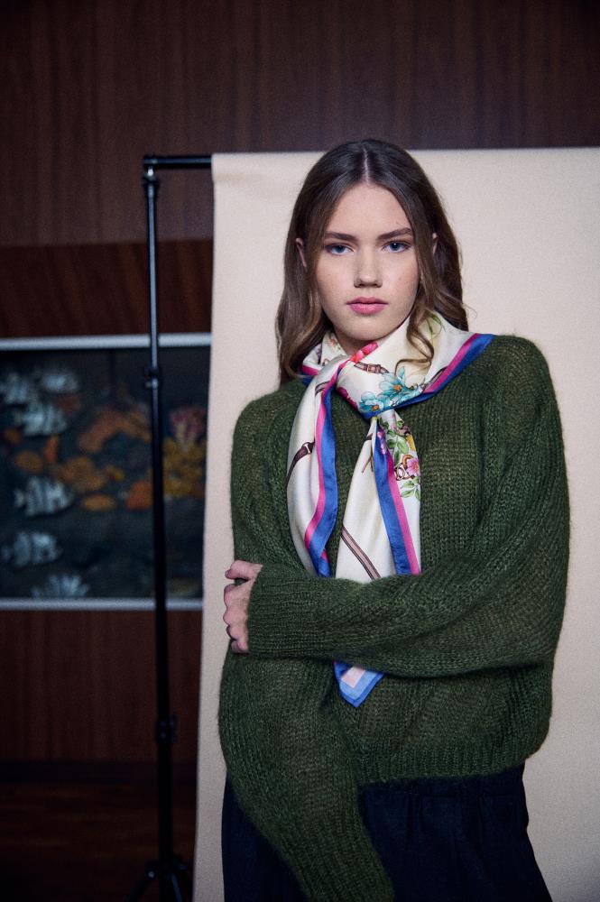 A young woman poses in front of a light studio backdrop and a section of a wooden wall. She is wearing a rich moss green, fluffy sweater, with a light silk scarf tied in a loose knot around her neck. The scarf is decorated with floral motifs and brown strap patterns, framed by bold pink and blue stripes along the edges. The model has a calm expression, and her pose with crossed arms creates a cozy and stylish spring or autumnal vibe.