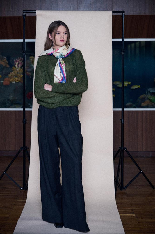 A person standing in front of a white backdrop wearing a green sweater and black trousers, with the silk scarf tied around the neck.