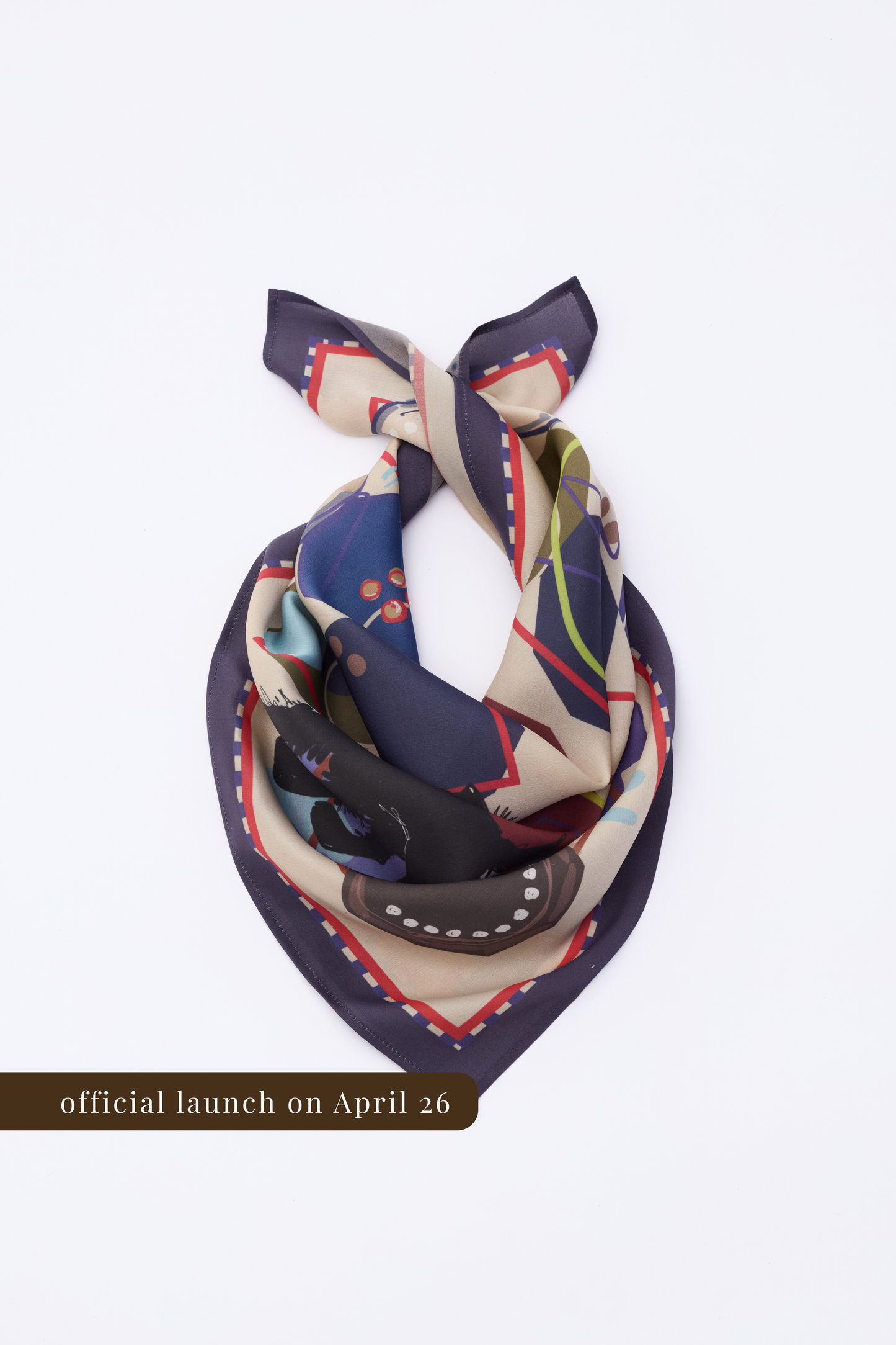 A square silk scarf featuring an abstract illustrative pattern, folded into a triangular shape on a white background. The design includes dark blue, cream, and olive green sections with red geometric borders. A brown banner at the bottom indicates the official launch date on April 26.