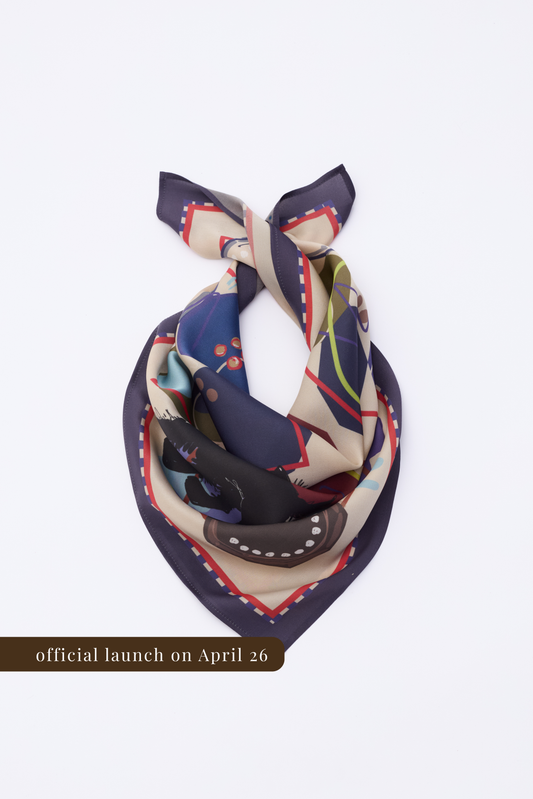 A square silk scarf featuring an abstract illustrative pattern, folded into a triangular shape on a white background. The design includes dark blue, cream, and olive green sections with red geometric borders. A brown banner at the bottom indicates the official launch date on April 26.