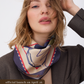 A close-up portrait of a model wearing a dark brown textured blazer and a silk scarf with a cream, navy, and red abstract pattern. The scarf is tied loosely around the neck, highlighting its satin finish. A brown banner at the bottom indicates the official launch on April 2