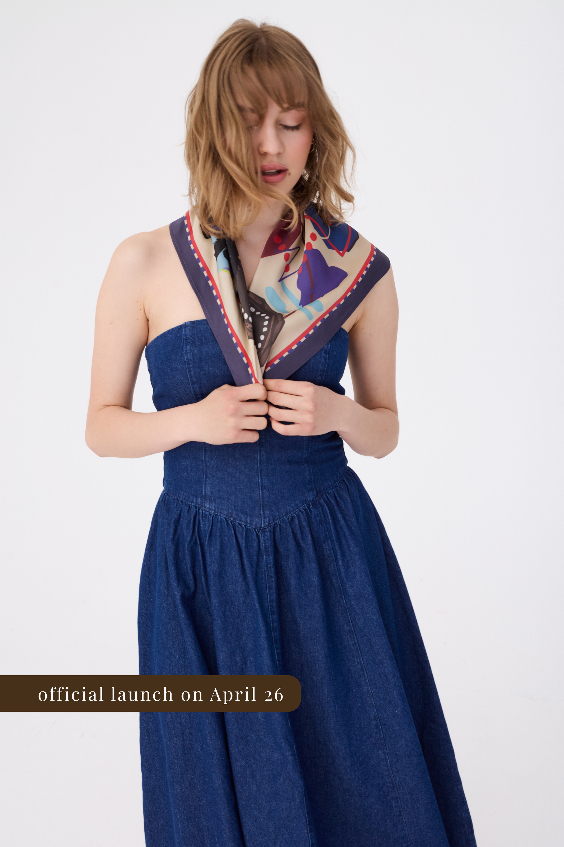 A woman wearing a strapless dark blue denim dress paired with a large silk scarf draped over her shoulders. The scarf features an abstract artistic print with purple, tan, and blue tones and a navy border. A brown banner at the bottom indicates the official launch on April 26.