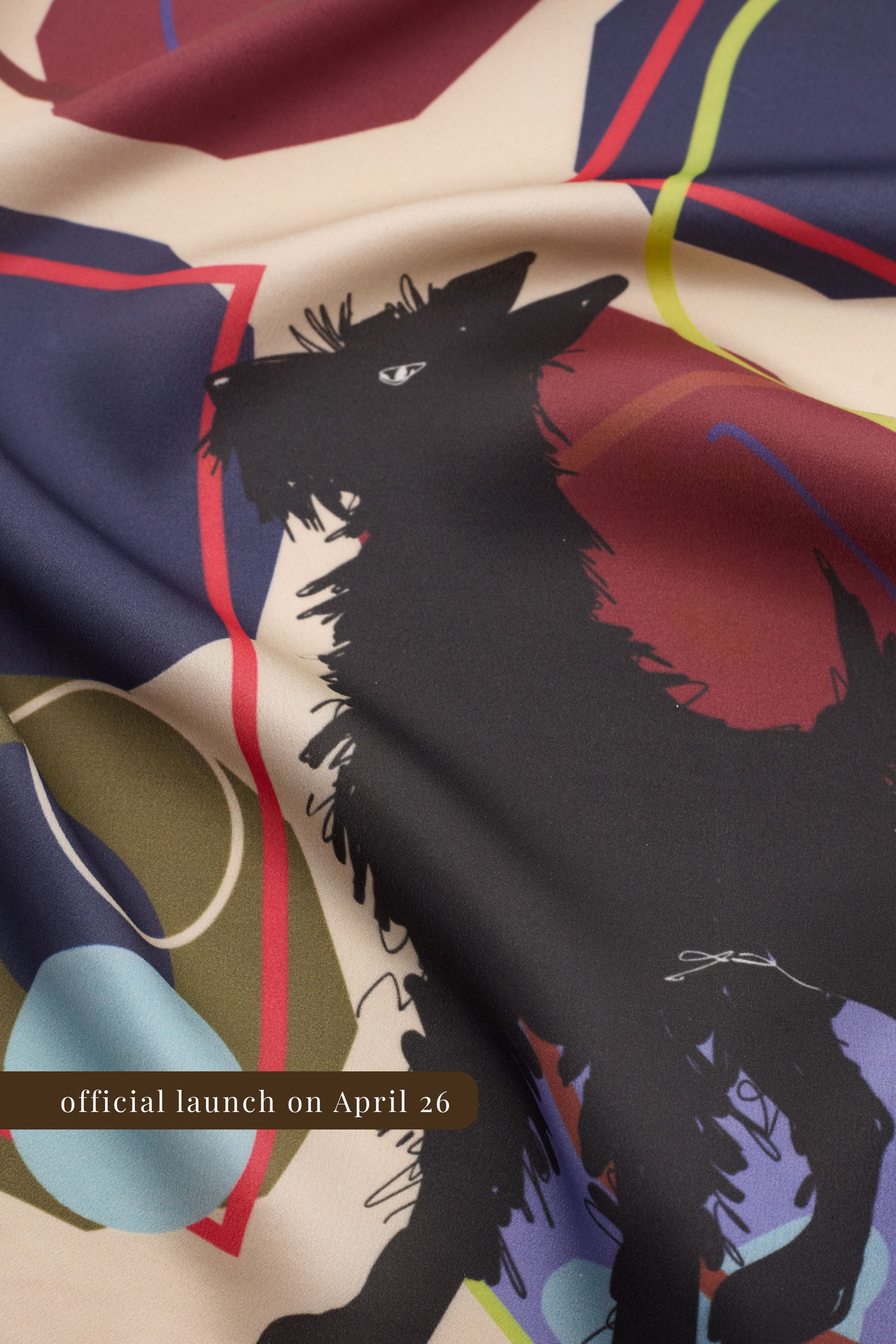 A macro shot of a silk scarf featuring a stylized black dog terrier illustration among abstract geometric shapes in burgundy, navy, and olive green. The fabric is draped to show its fine woven texture and soft matte finish. A brown banner at the bottom reads "official launch on April 26".