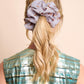 A back view of a woman with blonde wavy hair tied in a medium ponytail. The main focus is an oversized, voluminous greyish silk scrunchie featuring small ice cream patterns. She is wearing a shimmering blue and gold top against a light, neutral background.