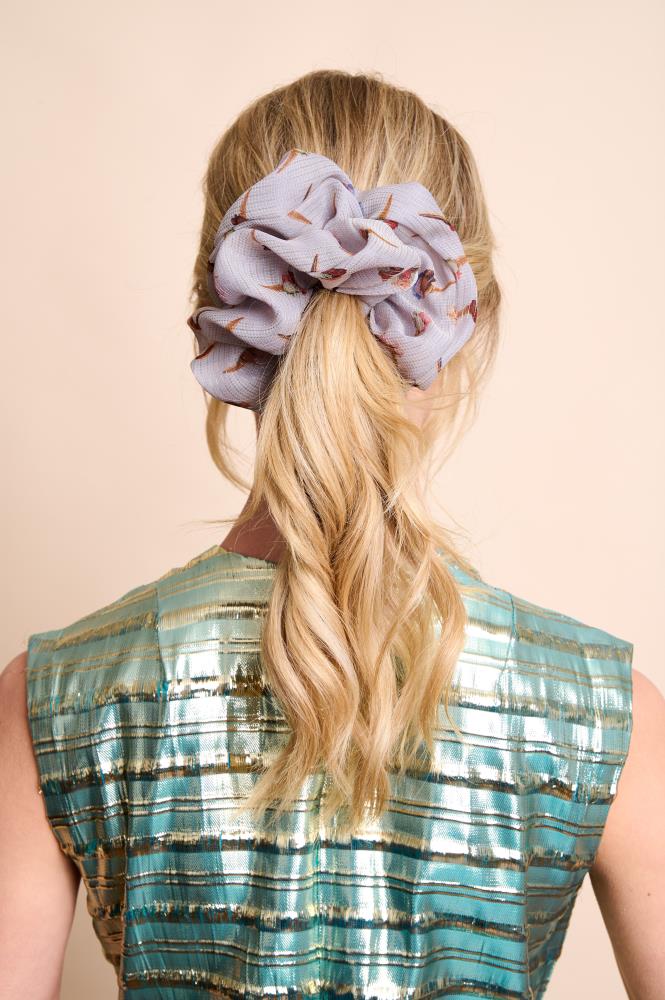 A back view of a woman with blonde wavy hair tied in a medium ponytail. The main focus is an oversized, voluminous greyish silk scrunchie featuring small ice cream patterns. She is wearing a shimmering blue and gold top against a light, neutral background.