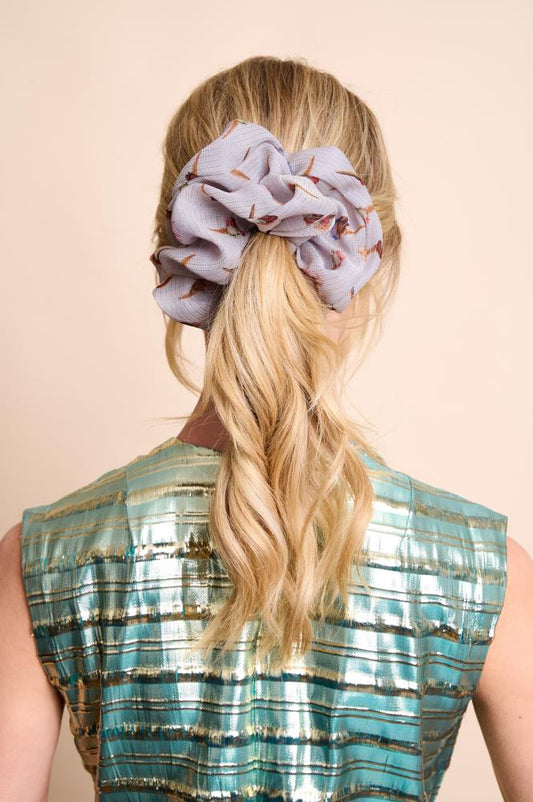 A back view of a woman with blonde wavy hair tied in a medium ponytail. The main focus is an oversized, voluminous greyish silk scrunchie featuring small ice cream patterns. She is wearing a shimmering blue and gold top against a light, neutral background.