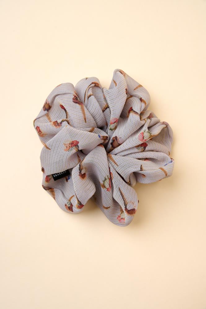 A large, voluminous fabric scrunchie placed against a warm yellowish background. The accessory is made from a greyish textured fabric featuring small ice cream patterns. A small black brand label is visible on the left side.