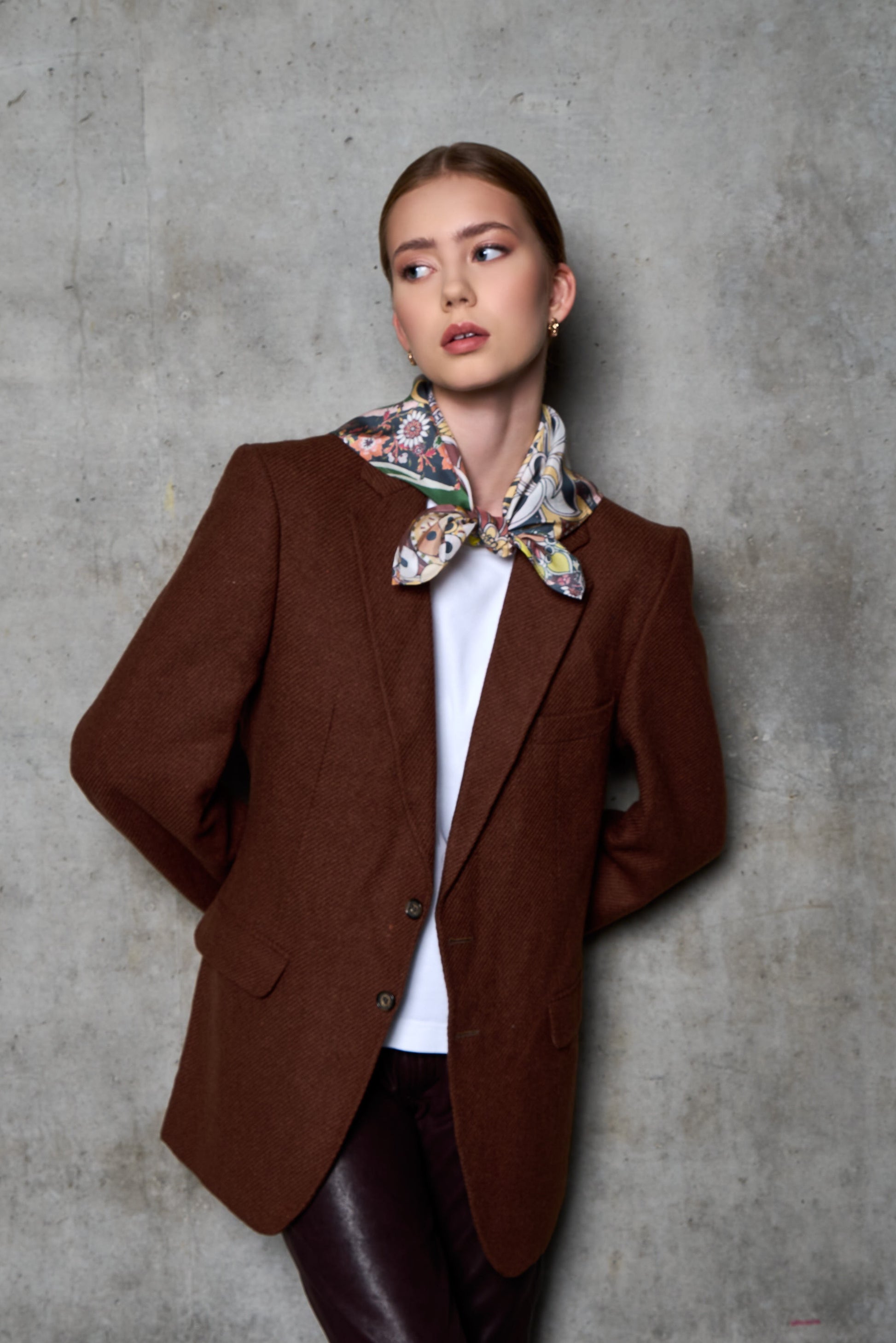 A woman poses against a grey concrete wall in an industrial setting, looking to the side. She is wearing a brown wool blazer, a white t-shirt, and dark burgundy leather trousers. Her look is accessorized with a vibrant, colorful silk scarf featuring floral and patterned motifs, stylishly tied in a knot at the front. Side lighting highlights the textures of the different fabrics – the coarse wool blazer, the smooth leather, and the shiny silk.