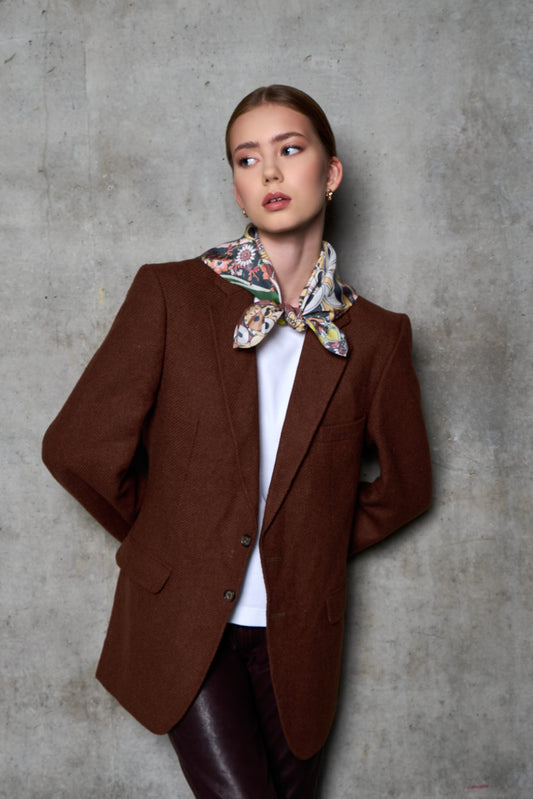A woman poses against a grey concrete wall in an industrial setting, looking to the side. She is wearing a brown wool blazer, a white t-shirt, and dark burgundy leather trousers. Her look is accessorized with a vibrant, colorful silk scarf featuring floral and patterned motifs, stylishly tied in a knot at the front. Side lighting highlights the textures of the different fabrics – the coarse wool blazer, the smooth leather, and the shiny silk.