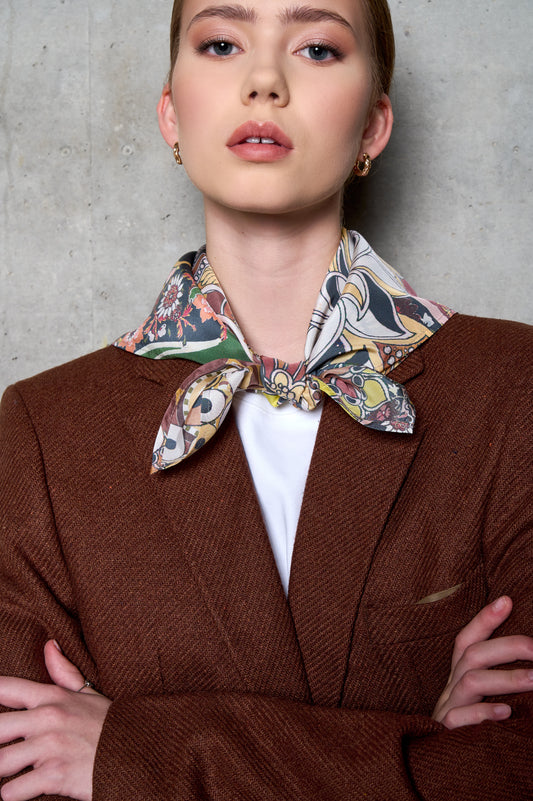 A close-up portrait of a woman posing against a concrete wall. She is wearing a textured brown wool blazer over a white t-shirt, with her arms crossed. A vibrant silk scarf featuring an intricate, colorful floral and paisley pattern is tied in a knot around her neck. The lighting highlights the fine texture of the blazer and the soft sheen of the scarf, creating a sophisticated and modern look.