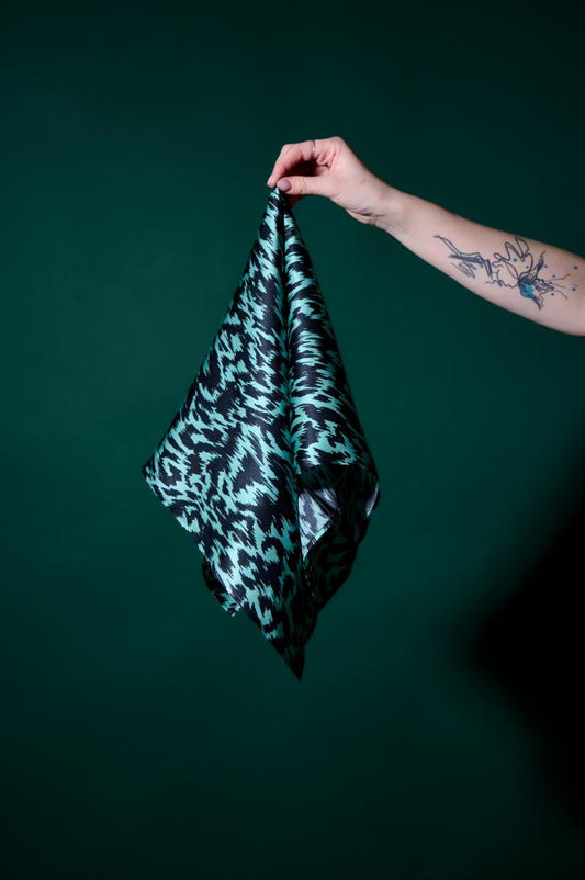 A hand with a tattoo holding a teal and black patterned silk scarf against a dark green background. The photo highlights the fabric's sheen, bold graphic pattern, and high-quality texture. 