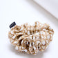 A large, voluminous fabric scrunchie placed on a light, neutral surface. The accessory features a cream-colored fabric with brown woven patterns, resembling wicker or basket weaving. A small black brand label is visible on the left side.