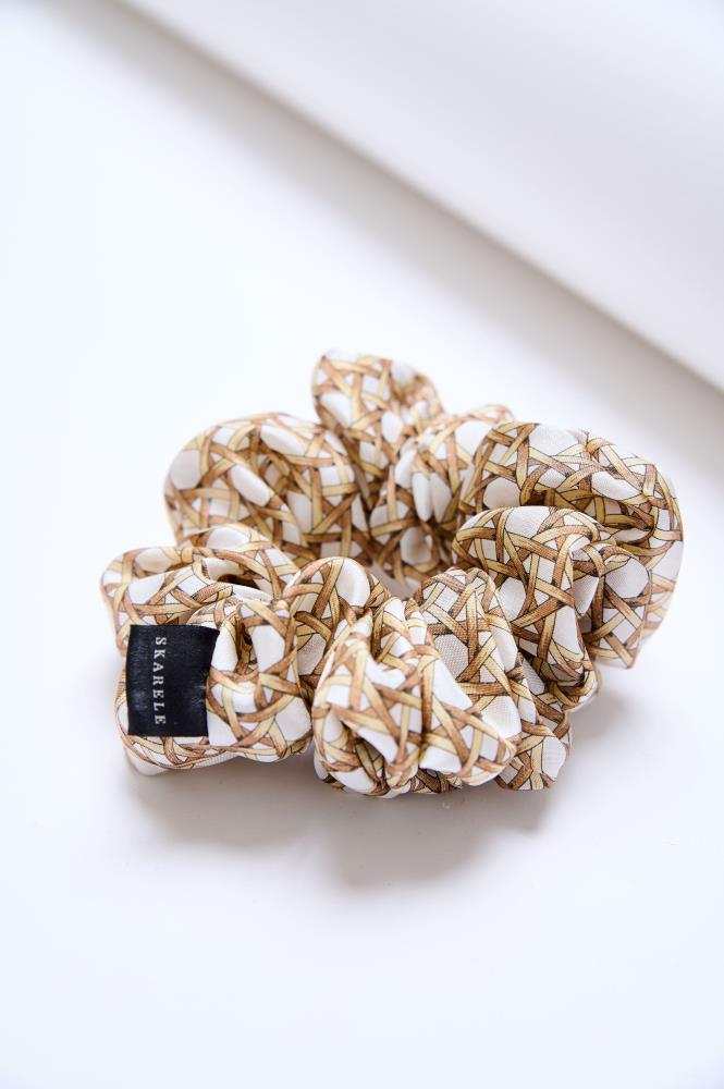 A large, voluminous fabric scrunchie placed on a light, neutral background. The accessory is made from cream-colored fabric featuring brown wicker-like woven patterns. A black label with the white text "SKARELE" is visible on the left side.
