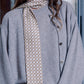 A woman poses wearing a grey sweater and a matching cardigan with dark buttons. A narrow, long silk scarf is wrapped around her neck, with the end hanging freely in front. The scarf features a small, repeating circular pattern on a brownish and cream background. The photo shows the model's profile, highlighting a subtle earring and an elegant, minimalist look.