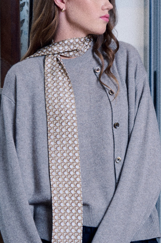 A woman poses wearing a grey sweater and a matching cardigan with dark buttons. A narrow, long silk scarf is wrapped around her neck, with the end hanging freely in front. The scarf features a small, repeating circular pattern on a brownish and cream background. The photo shows the model's profile, highlighting a subtle earring and an elegant, minimalist look.