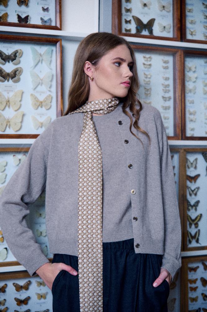 A young woman poses in front of a background of a science museum or a collector's room, featuring display cases with butterfly specimens. She is wearing a grey sweater and cardigan with dark trousers, and a narrow, long silk scarf is wrapped around her neck, with the end hanging freely in front. The scarf features a small geometric pattern. The model is looking to the side, and her calm pose combined with the unusual background creates an intellectual and stylish atmosphere.