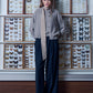 A full-length photo of a young woman posing in front of a wall lined with wooden display cases containing butterfly collections. She is wearing a grey sweater and cardigan with wide dark trousers. A narrow and very long silk scarf is stylishly wrapped around her neck, with the end hanging freely down almost to her waist. The scarf is decorated with a small geometric pattern. The photo features calm colors and a unique, scientific setting that highlights the elegant style of the outfit.