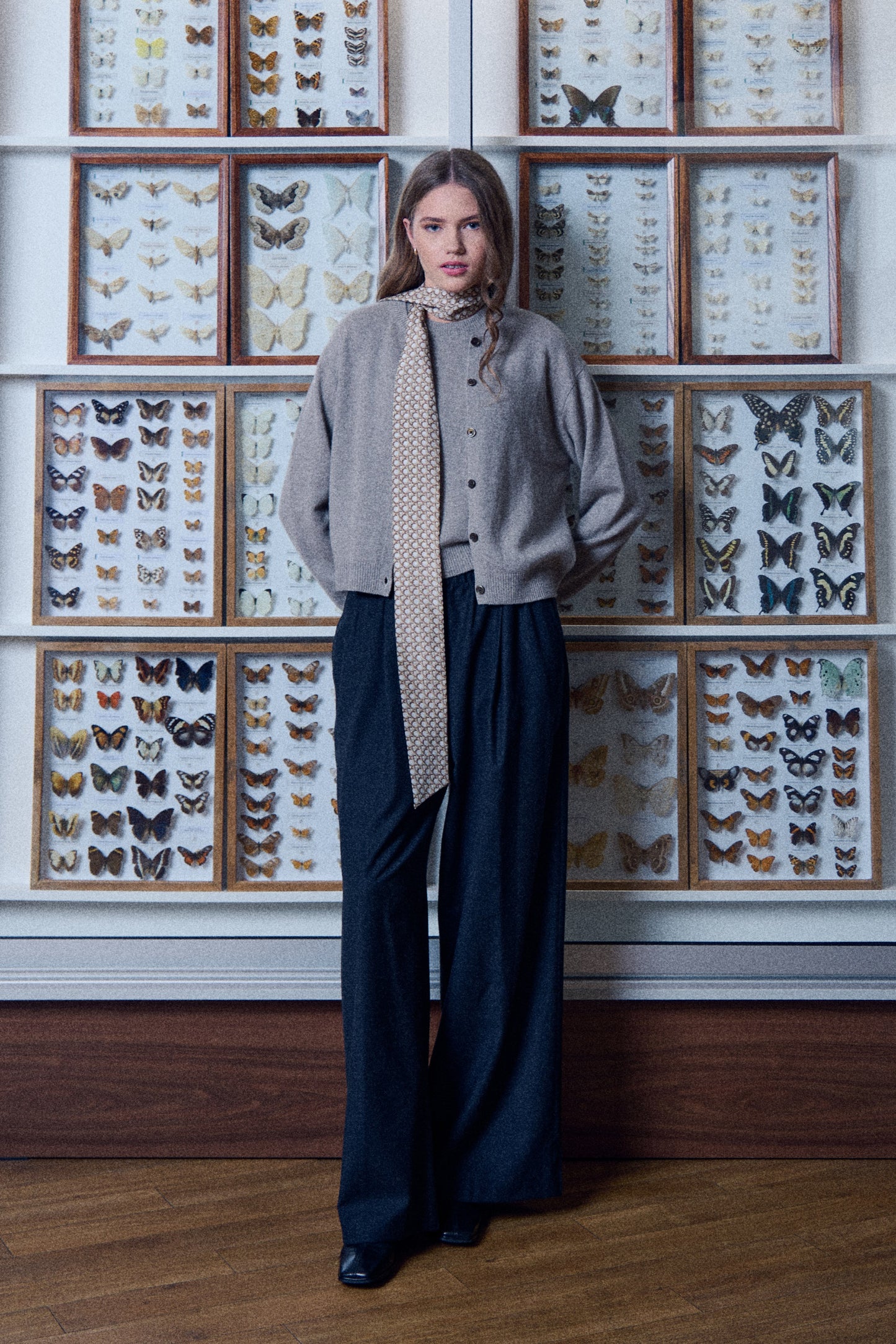 A full-length photo of a young woman posing in front of a wall lined with wooden display cases containing butterfly collections. She is wearing a grey sweater and cardigan with wide dark trousers. A narrow and very long silk scarf is stylishly wrapped around her neck, with the end hanging freely down almost to her waist. The scarf is decorated with a small geometric pattern. The photo features calm colors and a unique, scientific setting that highlights the elegant style of the outfit.