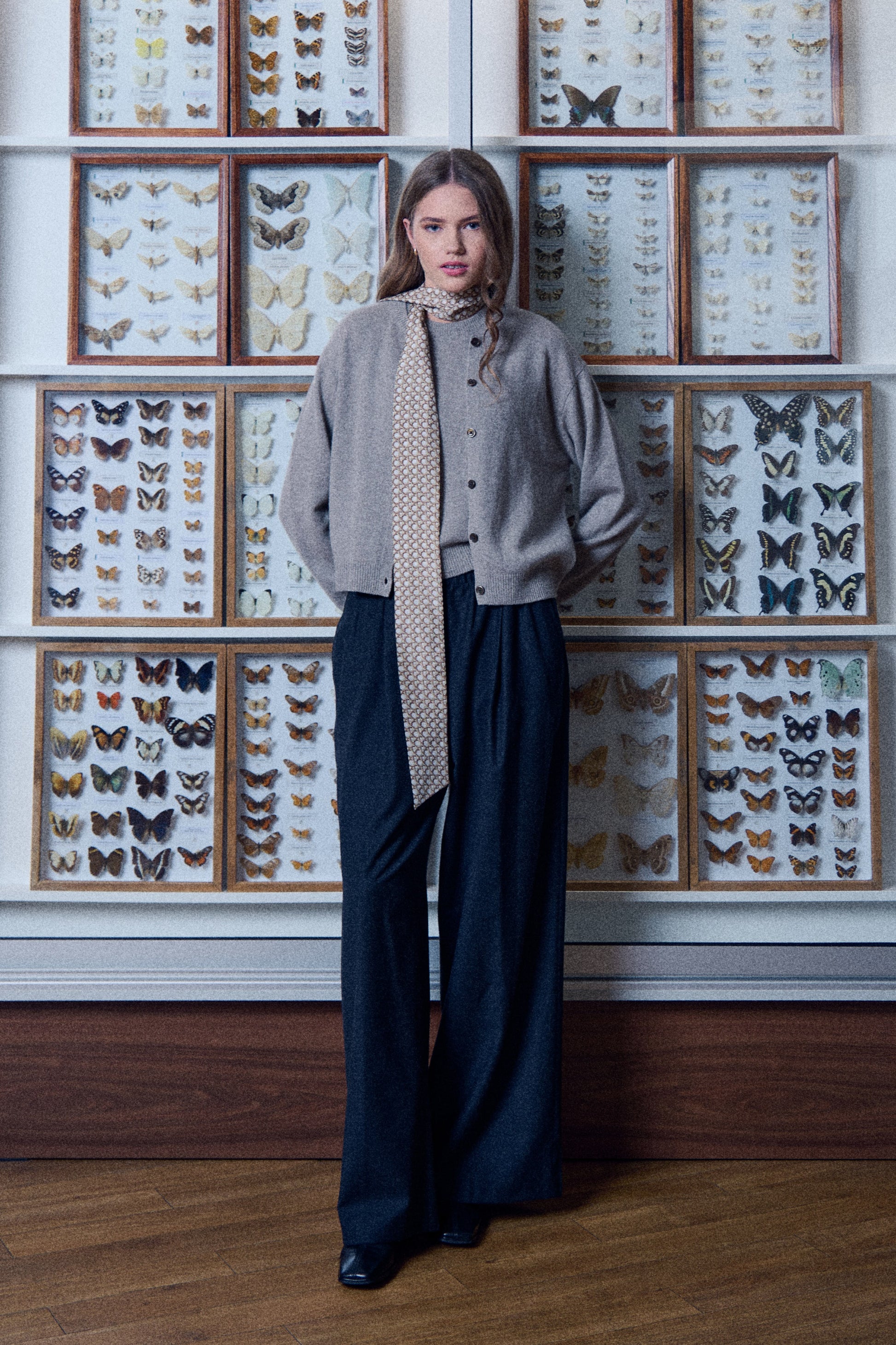A full-length photo of a young woman posing in front of a wall lined with wooden display cases containing butterfly collections. She is wearing a grey sweater and cardigan with wide dark trousers. A narrow and very long silk scarf is stylishly wrapped around her neck, with the end hanging freely down almost to her waist. The scarf is decorated with a small geometric pattern. The photo features calm colors and a unique, scientific setting that highlights the elegant style of the outfit.