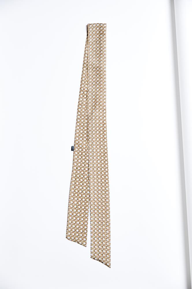 A narrow, long silk scarf (twilly style) stretched vertically against a white background. The accessory features a small, repeating geometric pattern resembling interwoven chains or a mesh motif on a brownish and cream background. The ends of the scarf are bias-cut, adding dynamism to the piece, and a small dark label is visible on the left side. The minimalist composition highlights the product's shape and subtle design.
