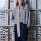 A model posing with a skinny silk scarf with a beige geometric pattern, paired with a taupe knit cardigan and dark high-waisted trousers, standing in front of framed butterfly collections.