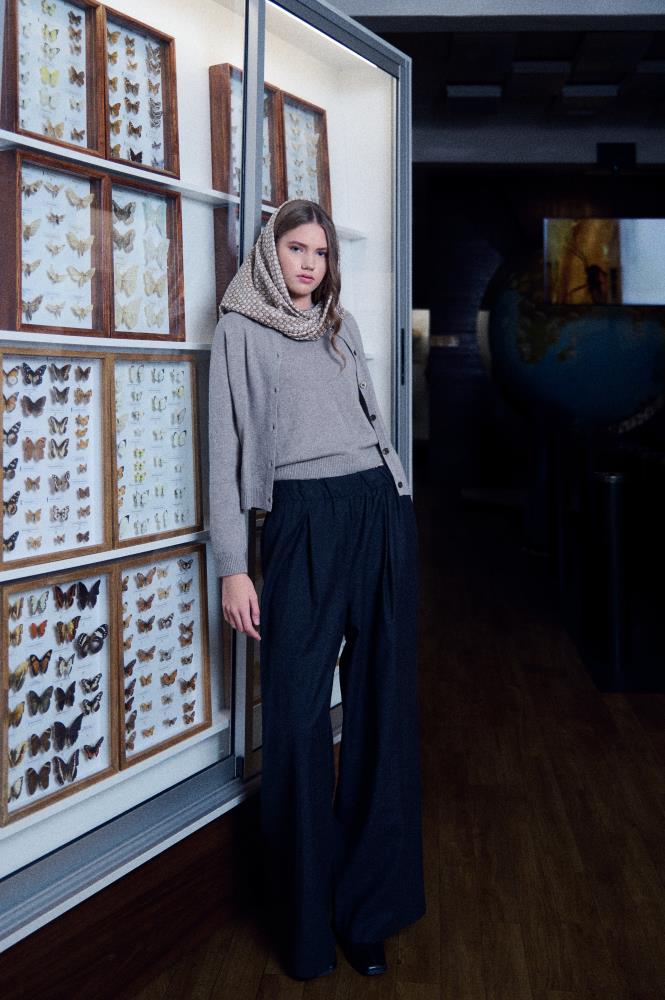 A full-length photo of a young woman posing in a dark museum interior, leaning against a lit glass display case containing a butterfly collection. She is wearing a grey sweater, cardigan, and wide dark trousers, with a light silk scarf featuring a fine geometric pattern wrapped around her head and neck. A large globe is visible in the distance, and the directional lighting highlights the model's silhouette and the textured pattern of the scarf, creating a mysterious and elegant atmosphere.