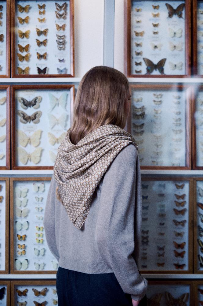 A back view photo of a young woman with light brown hair looking at a wall filled with butterfly collections in wooden display cases. She wears a grey sweater, with a silk scarf elegantly draped over her shoulders in a triangle shape. The scarf is decorated with a fine, textured geometric pattern on a brownish and cream background. The composition creates a calm, exploratory, and stylish mood, highlighting the accessory's texture in a scientific setting.