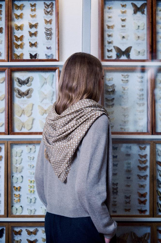 A back view photo of a young woman with light brown hair looking at a wall filled with butterfly collections in wooden display cases. She wears a grey sweater, with a silk scarf elegantly draped over her shoulders in a triangle shape. The scarf is decorated with a fine, textured geometric pattern on a brownish and cream background. The composition creates a calm, exploratory, and stylish mood, highlighting the accessory's texture in a scientific setting.