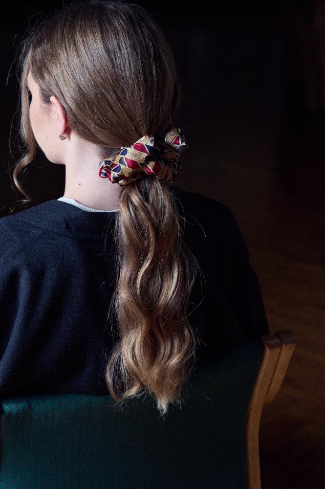 A person with long hair has their back turned to the camera, wearing a silk scrunchie in her hair. The scrunchie is visible and have a geometric pattern.