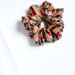 A large, voluminous fabric scrunchie placed on a clean white background. The accessory features a luxurious, detailed pattern with burgundy, gold, and navy blue motifs, reminiscent of classic ornamental style. The fabric's sheen and folds create a sense of depth and elegance.