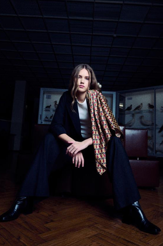 A young woman poses sitting on a leather bench in a dark, dramatic interior on wooden parquet floors. She is wearing a dark suit, a white t-shirt, and black boots, with a wide silk scarf stylishly draped over her left shoulder. The scarf features a dense geometric pattern with red, gold, and black elements. Glass display cases are visible in the background, and the low camera angle and lighting give the image a modern and edgy aesthetic.