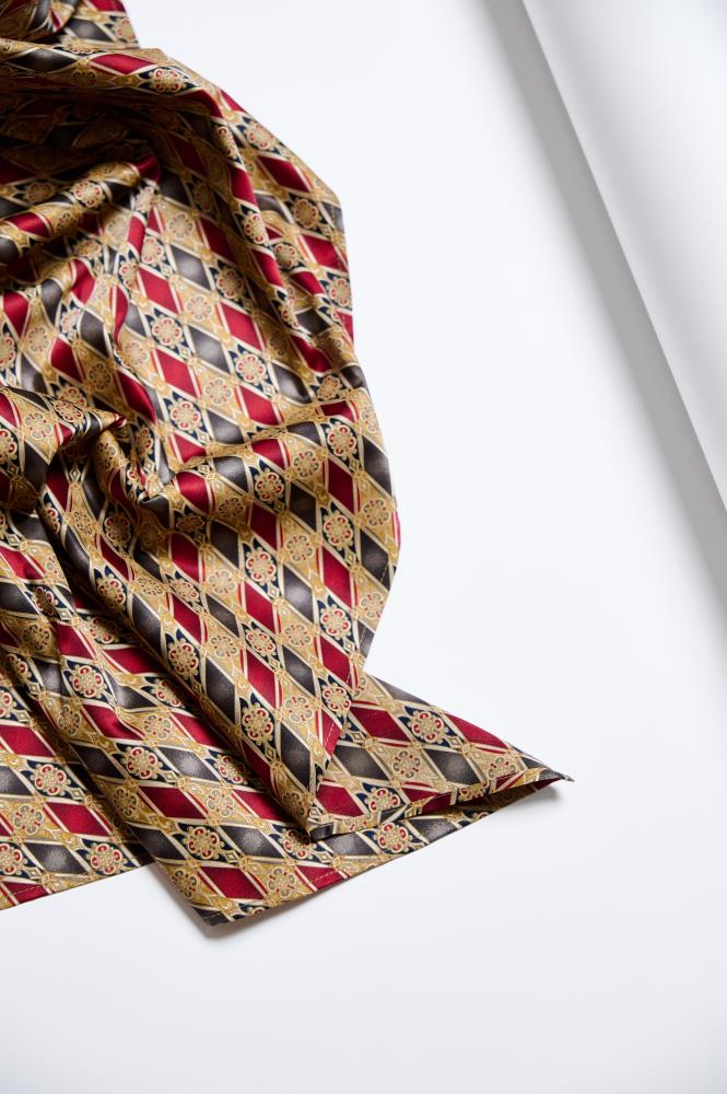An artistic composition against a light background featuring a luxurious, crumpled silk scarf. The fabric is adorned with a dense geometric pattern—red, gold, and black diamonds with small floral ornaments inside. The glossy surface of the natural silk and the deep folds emphasize the material's high quality and create an elegant, classic look.